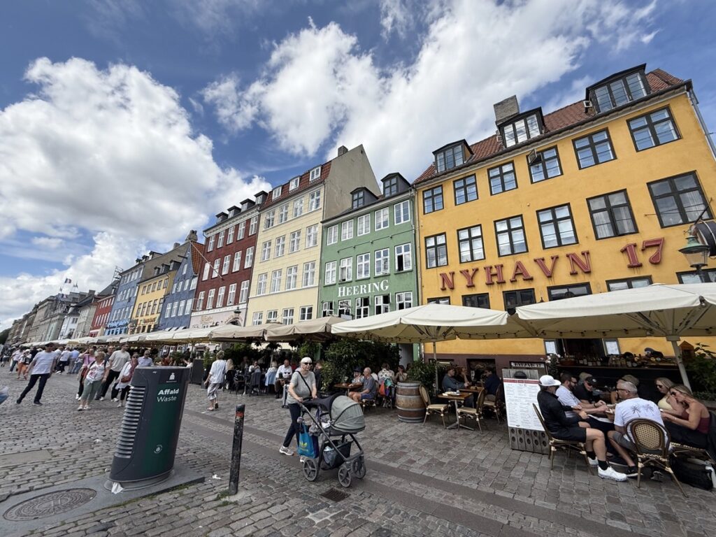 デンマーク・コペンハーゲンのNyhavn運河沿い。黄色・緑・赤のカラフルな建物と青空、テラス席に賑わう観光客（2024年夏）